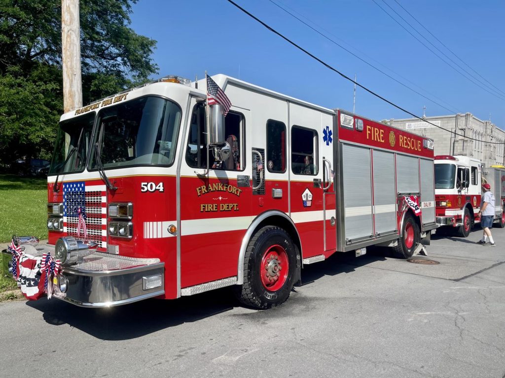 A red Frankfort fire truck with American flags and festive decorations is parked on a street. Another fire truck is visible in the background, and trees and a building are also in view.