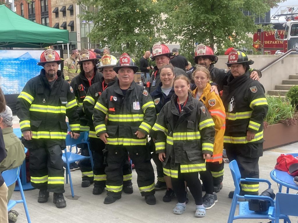 A group of firefighters in uniform, standing and posing for a photo outdoors. They are wearing helmets and reflective gear. Some greenery and a fire truck are visible in the background. They appear to be at an event or gathering.
