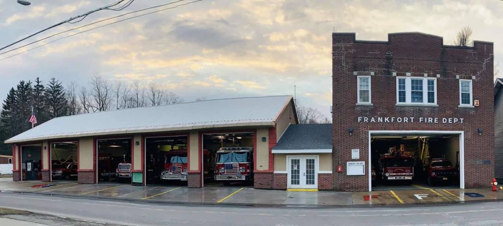 A two-story brick building labeled "Frankfort Fire Dept" with several garage bays containing fire trucks. The sky is cloudy, and trees are visible in the background.