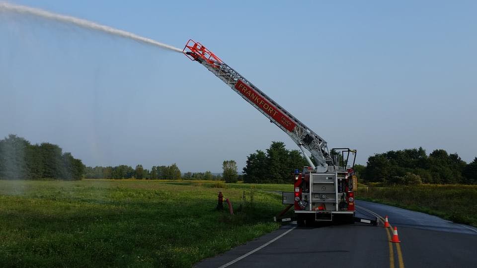 A fire truck extends its red ladder over a road, spraying water into the air. Cones are set up to guide traffic, and green fields stretch out on either side under a clear blue sky.